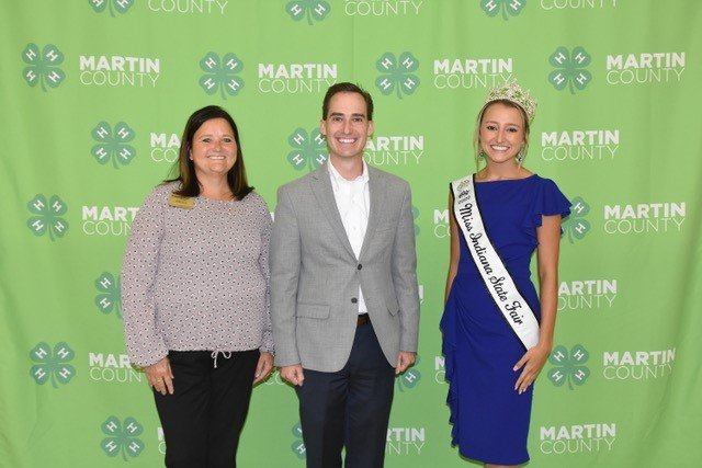 From left to right, women with brown hair stands next to a man with brown hair and a woman wearing a tiara.