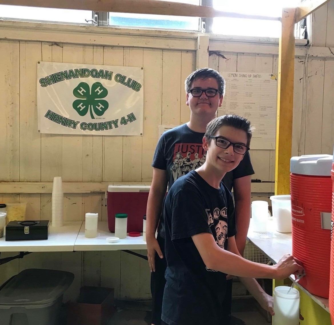 Two male youths stand filling a cup with lemonade in front of a Shenandoah 4-H club banner.