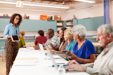 woman standing and holding a computer faces a row of elderly people working on laptops