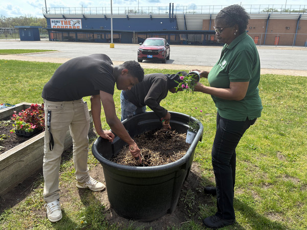 Image of Lake County 4-H Outreach Program Assistant holding flowers while two youth dig two holes in a raised garden bed outside in the grass. There are bleachers and a red vehicle behind them. 