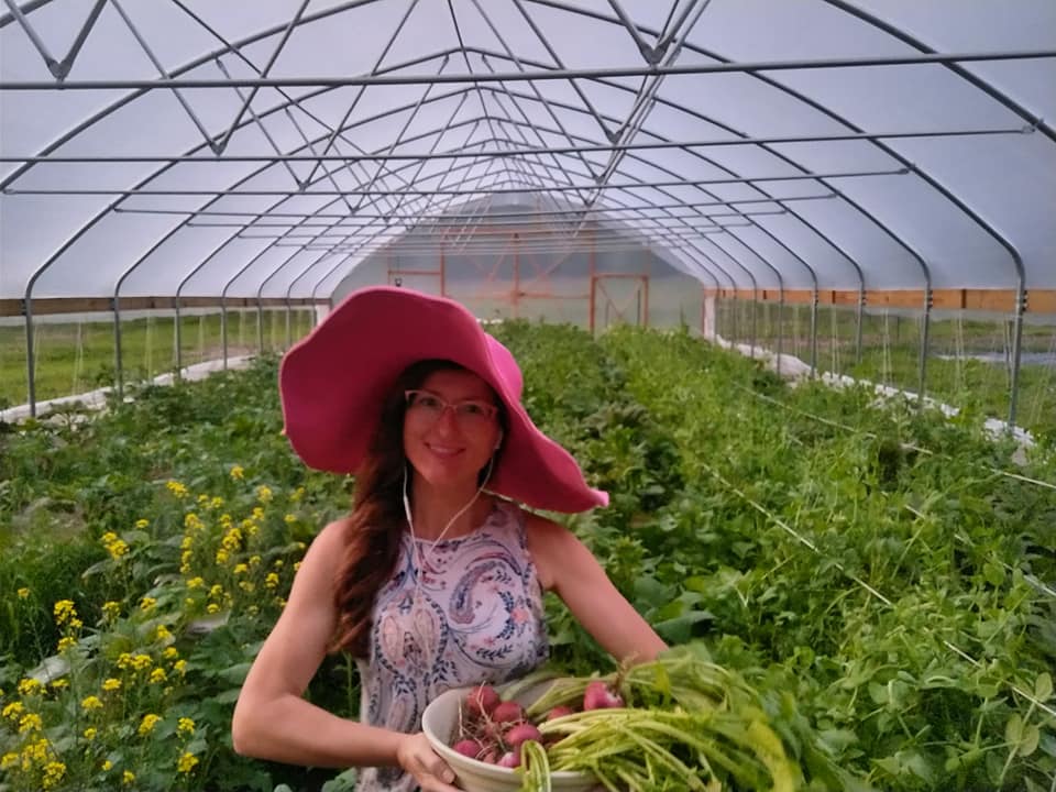 Woman stands in high tunnel greenhosue filled with plants wearing a pink hat and holding radishes.