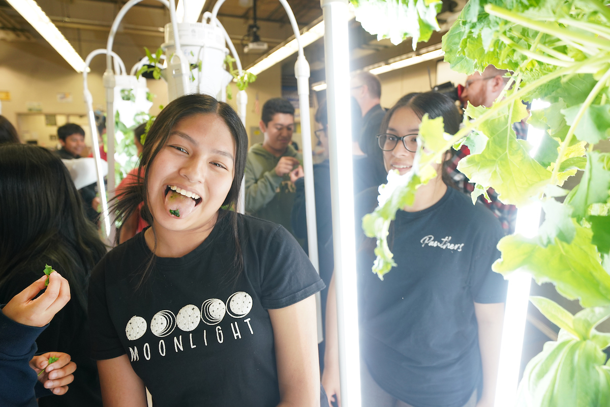 Teenage girl smiles while sticking her tongue out with leafy green on her tongue. Aeroponic towers and other youth are in the background.