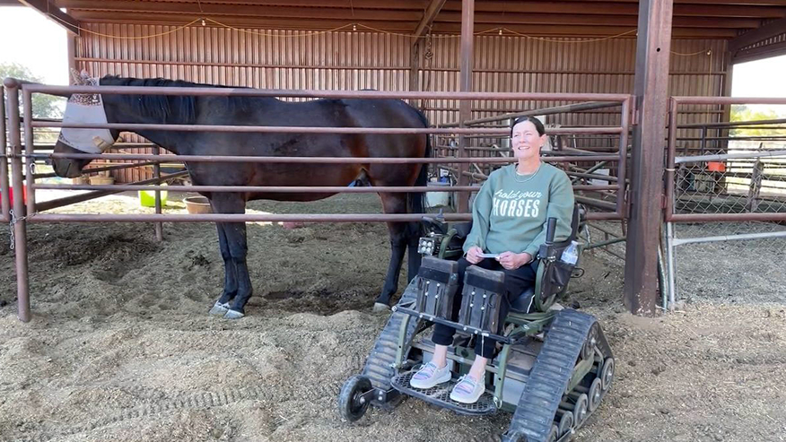 Tiffany Sanchez is seated in her specialized wheelchair beside a horse in a corral.