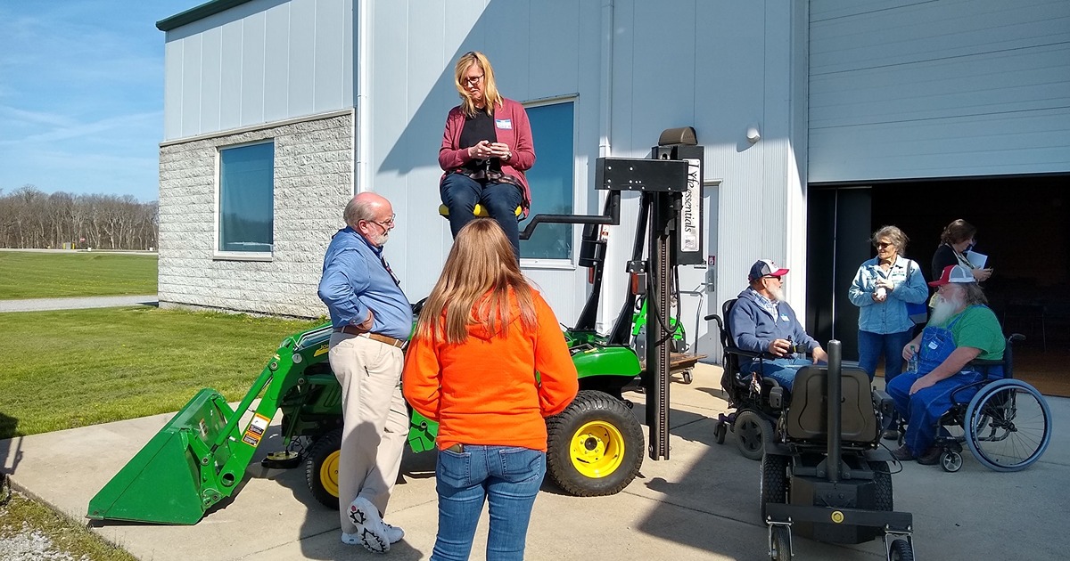 A group of event participants watches as a woman is lifted by a seat on a mechanical arm, which is attached to a small tractor. 