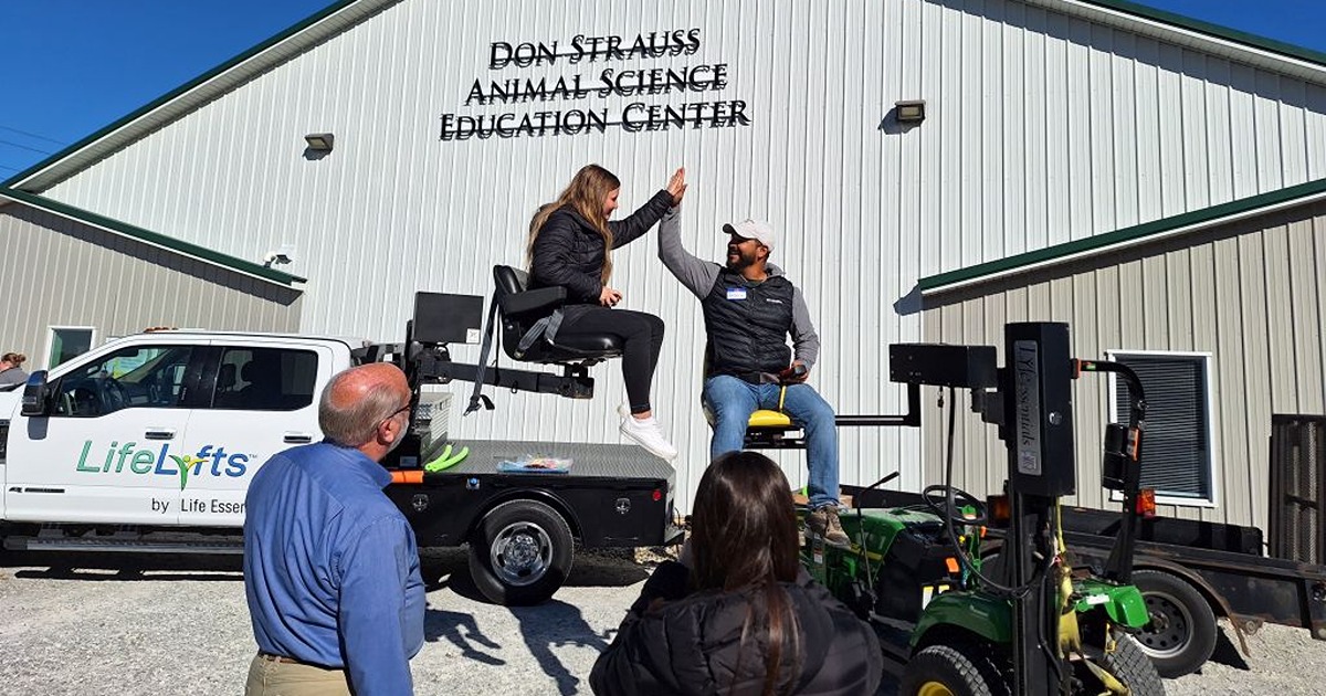 A man on a raised tractor seat high-fives a woman seated on a raised seat attached to a pick-up truck. 