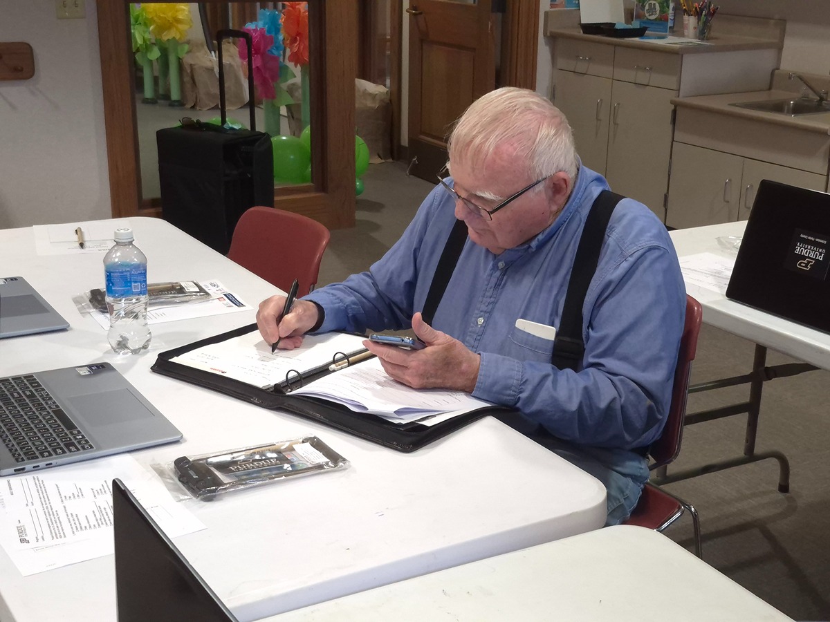 A man sits at a table. In one hand he holds his phone, and in the other hand he writes down notes onto a notepad.