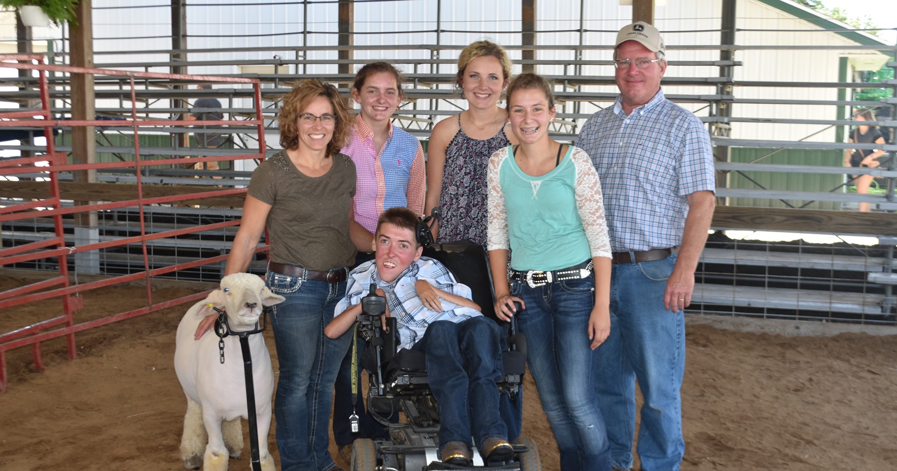 4-H volunteer Kristi Kretzmeier, left, stands with her husband, two daughters, two 4-H youth, and a show sheep. One youth, Kyle Albertson, sits in a motorized wheelchair, and his sheep is held in place[MCM1.1] by a specialized wheelchair attachment.