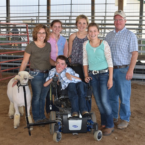 4-H volunteer Kristi Kretzmeier, left, stands with her husband, two daughters, two 4-H youth, and a show sheep. One youth, Kyle Albertson, sits in a motorized wheelchair, and his sheep is held in place[MCM1.1] by a specialized wheelchair attachment.