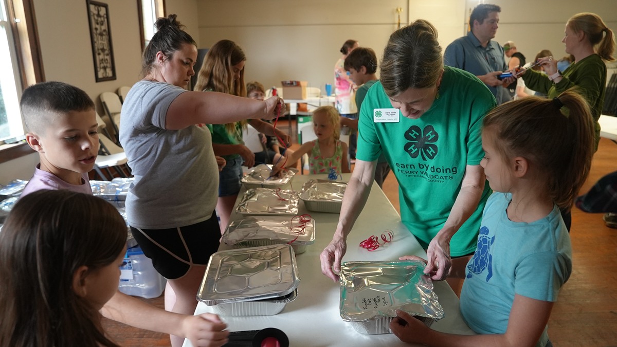 A 4-H volunteer shows youth how to seal the lid on an aluminum pan for the community project the group is working on.