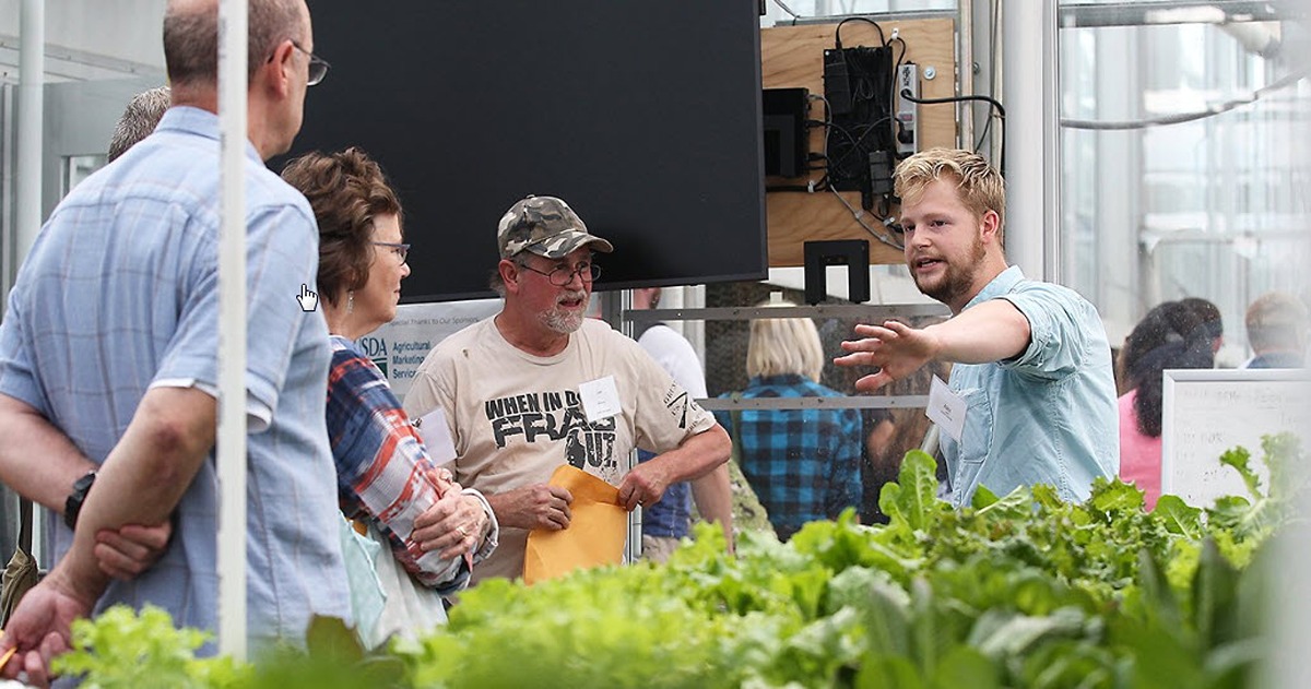Three participants, left, look at a presenter, right, who is gesturing to rows of hydroponically grown lettuce.